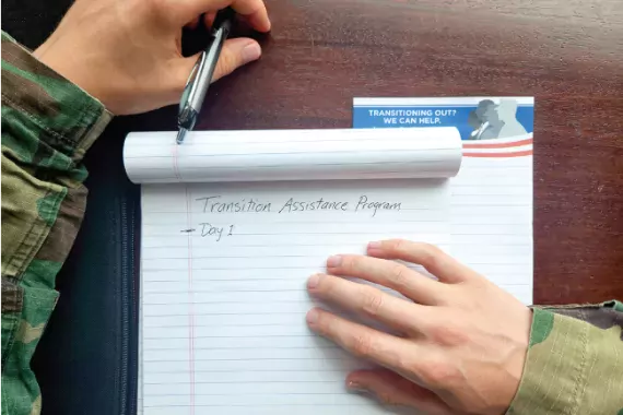 Stock art showing a person wearing military fatigues leaning over a legal notepad with the words &quot;Transition Assistance Program: Day 1&quot; written on it.